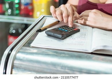 Hand Detail Of Latina Woman Using Her Small Calculator And Eco-friendly Cardboard Pen On A Large Notebook. Doing Calculations On Sales And Statistics In Her Grocery Store.