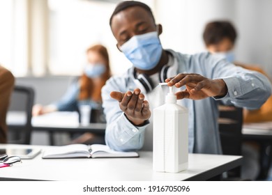 Hand Disinfection Concept. African american male student applying antibacterial sanitizer on hands from bottle, pouring it on palms, wearing protective medical mask, sitting at table at class - Powered by Shutterstock