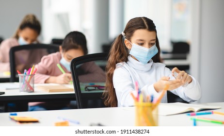 Hand Disinfection Concept. Small schoolgirl applying antibacterial sanitizer from small bottle, spraying it on palms, wearing disposable protective medical mask, sitting in classroom during lesson - Powered by Shutterstock