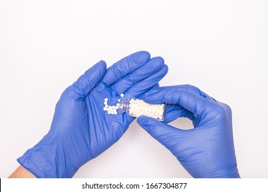 Hand Of A Doctor In Blue Latex Gloves Pours Homeopathic Globules Out Of A Glass Bottle Close Up On White Background, Alternative Medicine Concept.