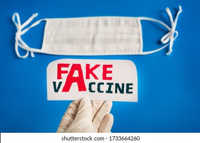 Hand Of A Doctor In Rubber Gloves Holds A Sheet With The Inscription Fake Vaccine. Medical White Mask On A Blue Background. Conspiracy Theory, Fake News Medicine.