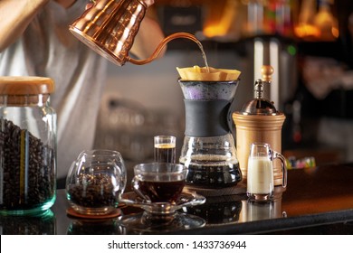 Hand Drip Coffee, Barista Making Drip Coffee By Pouring Spills Hot Water On Coffee Ground With Prepare Filter From Copper  Teapot To Glass Transparent Chrome Drip Maker. On Wooden Table Cafe Shop