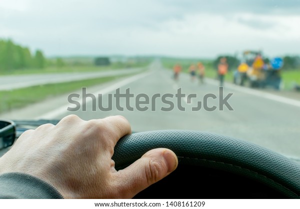 The hand of the\
driver who moves on a country road in cloudy weather in the evening\
on the car against the background of special equipment for repair\
of the road with warning\
signs