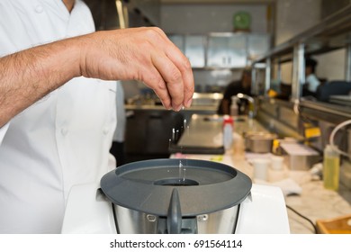 A Hand Dropping Organic Fresh Rock Salt Into A Food Blending Processor, In An Industrial Kitchen Setting.