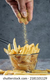 Hand Dropping Some Salt On A Basket Full Of French Fries On A Gray Surface And Gray Background