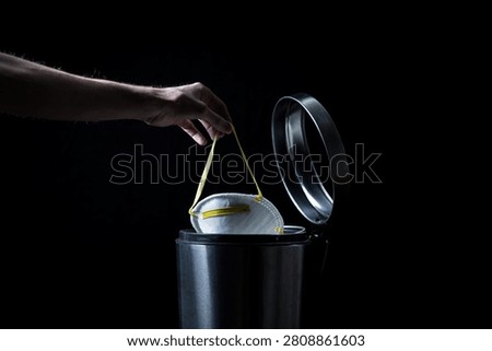 A hand drops a used protective face mask into an open stainless steel trash bin against a dark backdrop.