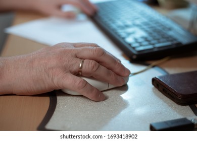 Hand Of An Elderly Man Lies On A Computer Mouse, At A Computer Table, Retired At Work At Home