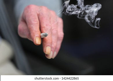 The Hand Of An Elderly Person, Holding A Lit Up Cigarette. The Nails Of His Index Finger And Ring Finger Are Completely Turned Yellow Because Of The Tar, Nicotine And Smoke. Concept Of Chain Smoker.