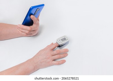 The Hand Of An Elderly Woman Holds A Mobile Phone Or Smartphone, Measures The Saturation And Oxygen Level In The Blood With A Modern Medical Device Pulse Oximeter On A White Background.