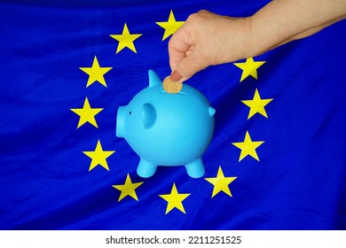 Hand Of Elderly Woman Putting Coin Into Piggy Bank On EU Flag Background. Hand Putting Coin To Piggy Bank. Retirement Savings, Piggy Bank. Concept Saving Money, Investment And Retirement Fund In EU