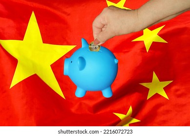 Hand Of Elderly Woman Putting Coin Into Piggy Bank On China Flag Background. Hand Putting Coin To Piggy Bank. Retirement Savings, Piggy Bank. Concept Saving Money And Retirement Fund In China