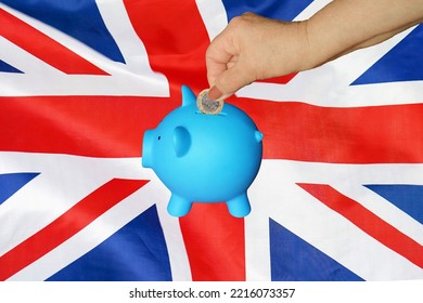 Hand Of Elderly Woman Putting Coin Into Piggy Bank On UK Flag Background. Hand Putting Coin To Piggy Bank. Retirement. Concept Saving Money And Retirement Fund In United Kingdom Of Great Britain