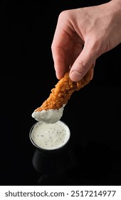 A hand elegantly dips a crispy chicken tender into a rich and creamy sauce, highlighting a delightful contrast against the sleek black background. A moment of indulgence captured.