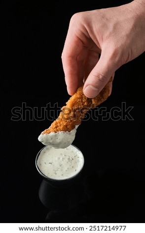 A hand elegantly dips a crispy chicken tender into a rich and creamy sauce, highlighting a delightful contrast against the sleek black background. A moment of indulgence captured.