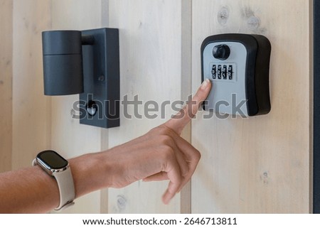 Hand entering code on outdoor key lock box. Close-up of a person inputting a combination code into a secure wall-mounted key lock box near a modern door and light fixture.