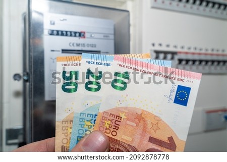 Hand with euro banknotes in front of an electricity meter as electricity is very expensive because of the energy crisis