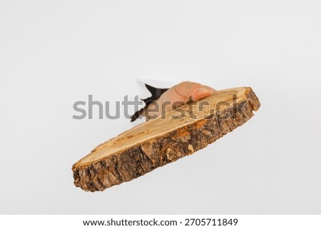 A hand extends through a hole in a plain wall, gripping a rustic wood slice. The unique scene highlights the contrast between natural and modern elements.