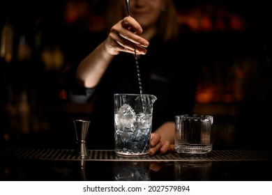 hand of female bartender holds spoon and stirred cold cocktail in crystal mixing cup. Clear old-fashioned glass and steel jigger stand side by side on the bar