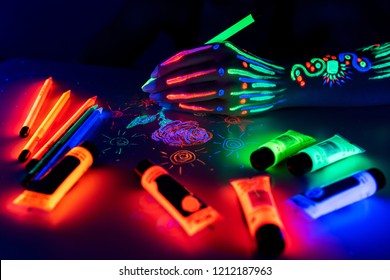 Hand with fluorescent ethnic pattern holding neon pencil. Fluorescent paint and pencil are on the table. Body Art concept