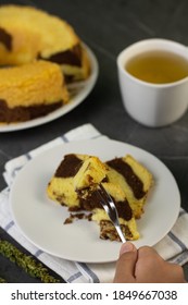 Hand In Frame Slicing Of Marmer Cake Or Marbel Cake On White Ceramic Plate With Fork, Served With A Cup Of Tea. Black Marble Background. 