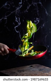 Hand In Frying Pan Making Fresh Vegetables Fly Through The Air Sautéing With Liquid On Black Background