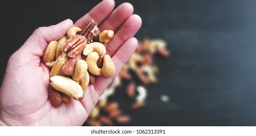 Hand Full of Mixed Nuts Over More Nuts in the Background on Black Table. Selective Focus on Hand. Top View. 