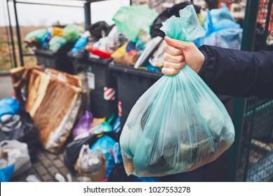 Hand with garbage against full trash cans with rubbish bags overflowing onto the pavement. - Shutterstock ID 1027553278