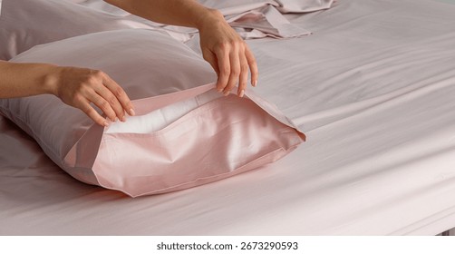 A hand gently pulls back a silky pillow cover in a soft pink bedding arrangement, creating an inviting atmosphere in a modern bedroom setting during daylight.