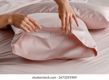 A hand gently pulls back a silky pillow cover in a soft pink bedding arrangement, creating an inviting atmosphere in a modern bedroom setting during daylight.