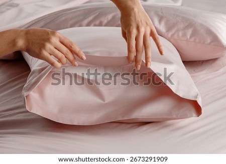 A hand gently pulls back a silky pillow cover in a soft pink bedding arrangement, creating an inviting atmosphere in a modern bedroom setting during daylight.