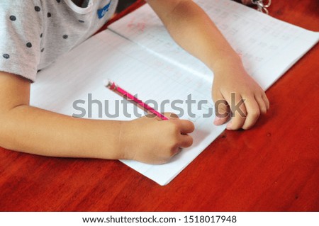 Hand of girl or kid holding Pencil and doing her homework by writing in a book on wood Table in Home. She is writing the word Crow in Thai.