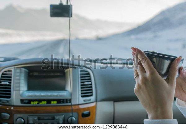 the hand of the girl in the\
passenger seat of the car holding hot tea from the mug from the\
thermos, view of the mountain winter road outside the car\
window