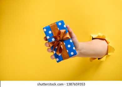 Hand Giving A Gift Box Through Torn Yellow Paper Wall. Polka Dot Package And Satin Ribbon Bow. Special Offer Or Sale Concept.