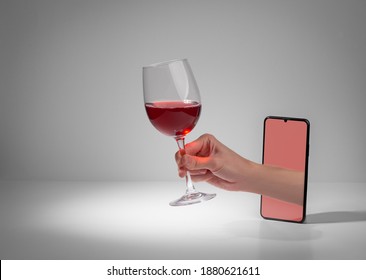Hand giving a glass of red wine from out of a phone screen. Online wine market service and party at home during a quarantine.