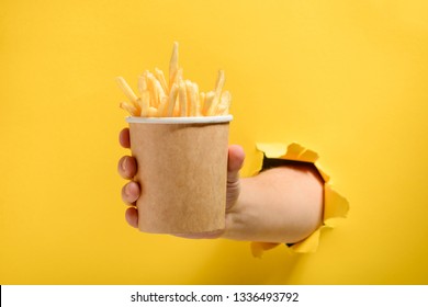 Hand Giving A Paper Cup Of French Fries Through Torn Yellow Paper. Special Offer In A Fast Food Restaurant.