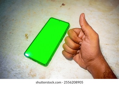 A hand giving a thumbs up gesture next to a smartphone with a bright green screen on a white surface, symbolizing approval or satisfaction with the device - Powered by Shutterstock