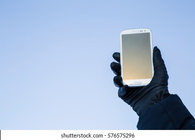 Hand In The Glove Holding A Mobile Phone On The Blue Sky Background.