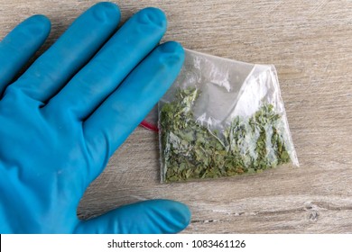 Hand In The Glove Presses To The Table A Bag Of Synthetic Smoking Substance, Cannabis, Weed, Marijuana