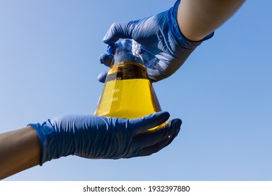 Hand with gloves holding beaker with ethanol biofuel against blue sky, observing its purity.