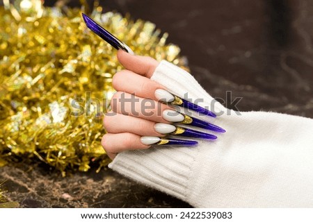A hand gracefully presents a set of long, white and purple glitter nails with elegant gold foil accents, complemented by a sparkling tinsel backdrop