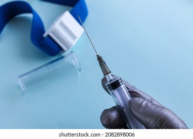 A hand in a gray medical glove holds a syringe, a test tube and a tourniquet for taking blood from a vein are lying next to it. light blue background, close-up.