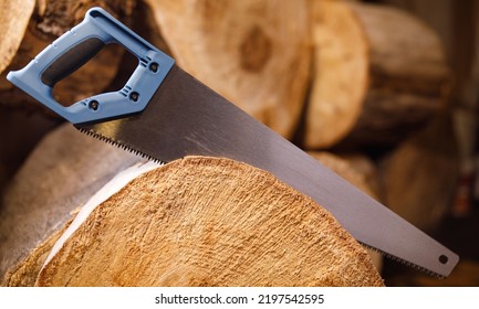 A Hand Hacksaw With A Blue Wood Handle Saws A Log. The Background Is Made Of Wood Logs.