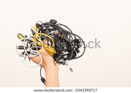 Hand held up with pile of tangled old smart technology wires, used charging cables and cords. E-waste, planned obsolescence, electronic donation, disposal of electronic waste, recycling concept
