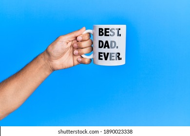 Hand Of Hispanic Man Holding Best Dad Ever Coffee Cup Over Isolated Blue Background.