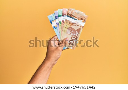 Hand of hispanic man holding canadian dollars over isolated yellow background.
