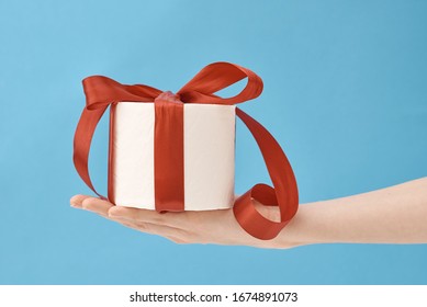 Hand Hold Toilet Paper Wrapped In A Festive Ribbon As Gift On Blue Background