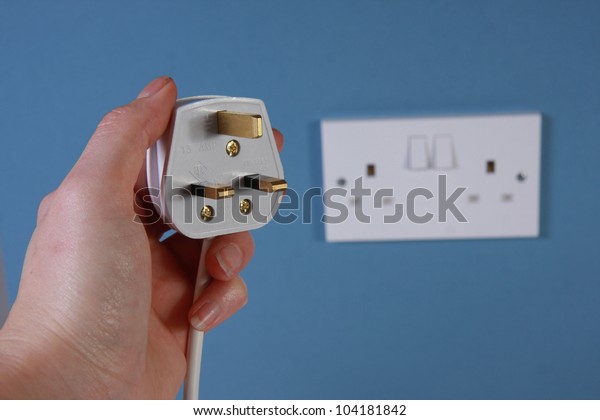 Hand holding a 3 pin\
plug with socket in the background on a blue wall.  This is a\
United Kingdom 3 pin plug