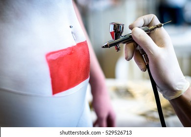 Hand Holding Air Brush In Body Painting Woman Body. Paintbrush Artist Is Covering Body With White And Red Paint Using A Template To Make A Tatoo.