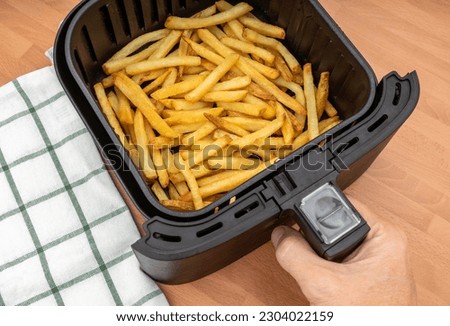 HAND HOLDING AIR FRYER BASKET WITH FRESHLY MADE FRENCH FRIES OR FRIED POTATOES IN THE KITCHEN. ULTRA PROCESSED VEGAN FOOD. TOP VIEW.
