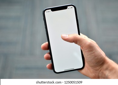 A hand is holding a Apple iPhone X on a plane background. iPhone X has a blank white background on its screen. The picture is shot in Istanbul,Turkey on 24/11/2018
